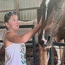 Girl in white tank top with brown hair in pony tail pets the head of a brown horse